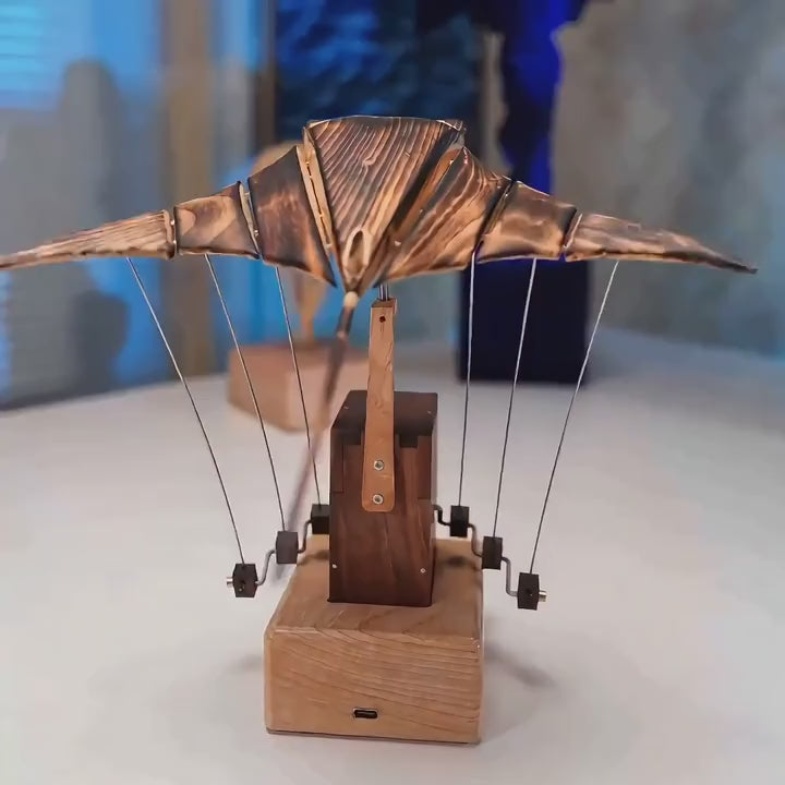 Front view of the manta ray automaton, copper-toned ribbed wings spread on wires above a dark wood tower and light wood base.
