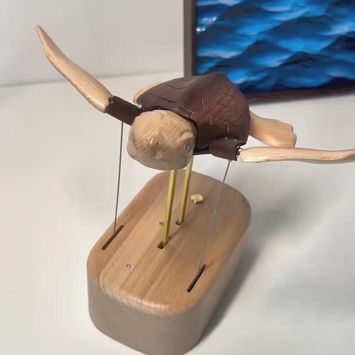 Sea turtle automaton on its oval wood base held up by brass rods, flippers outstretched, a blue sea painting behind.