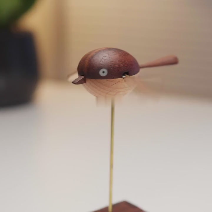 The round wooden bird beats its wings fast on the brass rod above its dark wood box, seen from the side, the front and three-quarter.