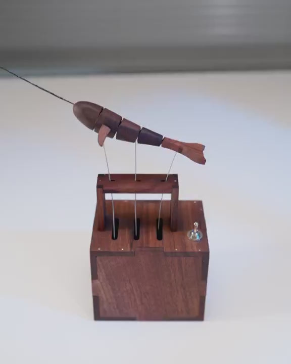 Segmented dark narwhal automaton swimming in place above its dark wood box on a white table.