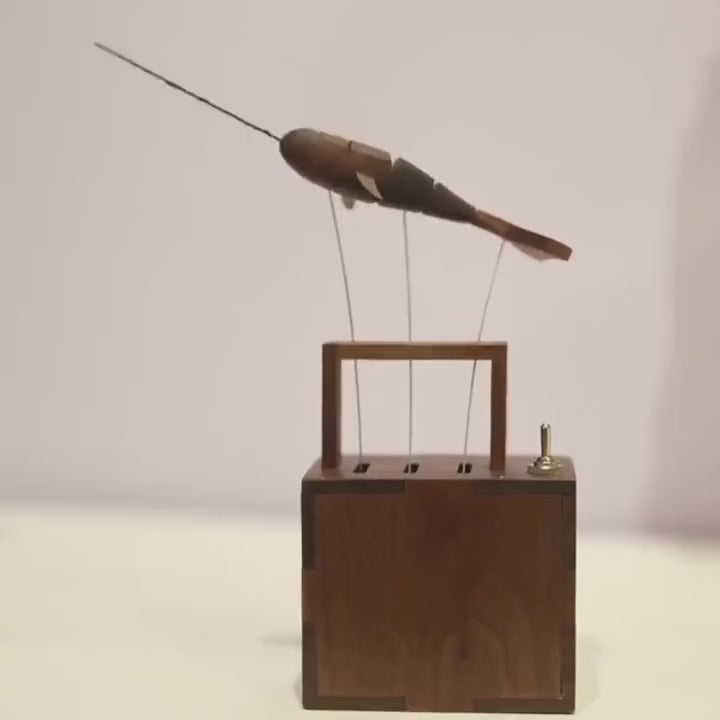 dark wood whale on three wires above a slotted dark wood box with a small toggle switch on top, tail whipping up and down, first against a pale wall and then on a desk.