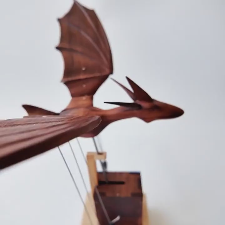 A dark walnut dragon with jointed wings glides on fine wires above a small walnut box on a light wood base, wings rising and falling, white background.