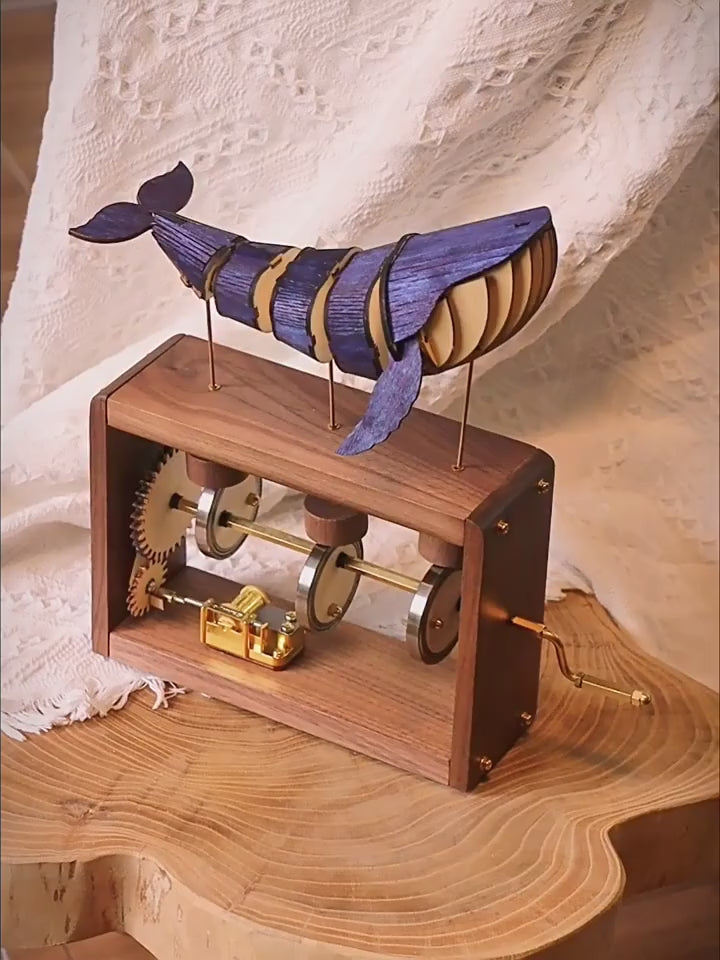 A hand turns the brass crank on a walnut music box; a blue-violet jointed wooden whale rolls on three brass rods above, gears and cams turning inside the open frame.