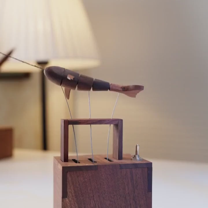 dark wood whale with a white fin swinging through the air on its wires, a pleated lamp glowing behind and a dovetailed dark wood frame below.