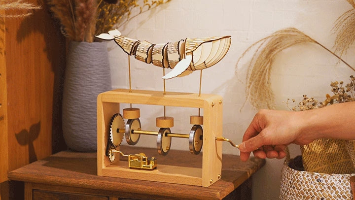 A hand turns the crank on the beech edition and the pale whale rolls on its rods, a vase of dried grass behind.