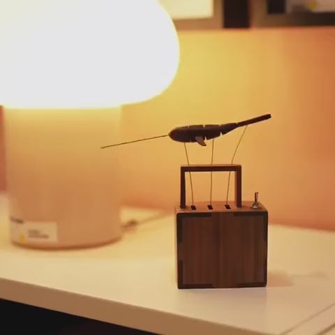 Silhouette-like side view of the dark wood whale on wires above its square box, a bright orange lamp glowing behind.