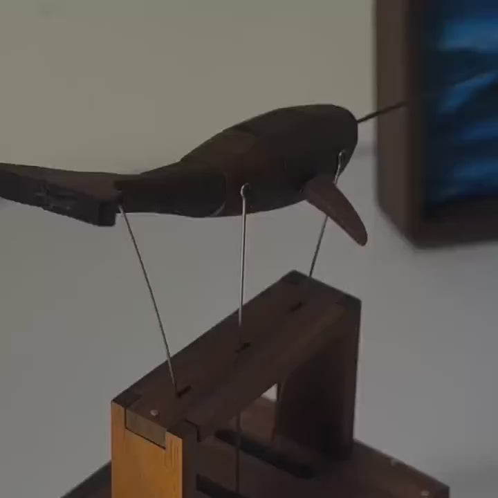 Dark narwhal automaton with a long tusk above its dark wood frame, a blue sea painting behind, ending close on the tail.