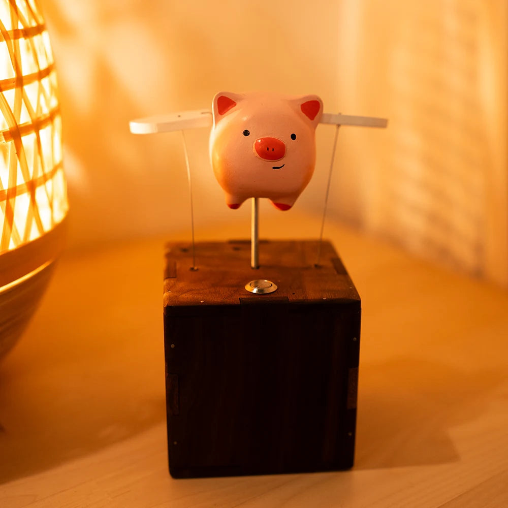 Pink pig with white wings on thin wires above a dark wood box with a small metal button on top, a glowing woven lamp and a warm wall behind.
