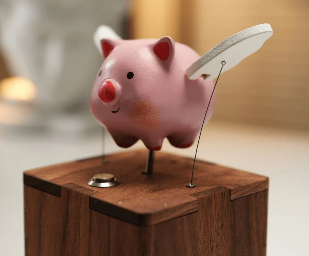 Close three-quarter view of the pink pig with a white wing raised on a thin wire above the finger-jointed corner of a wood box with a metal button, a blurred plaster bust behind.
