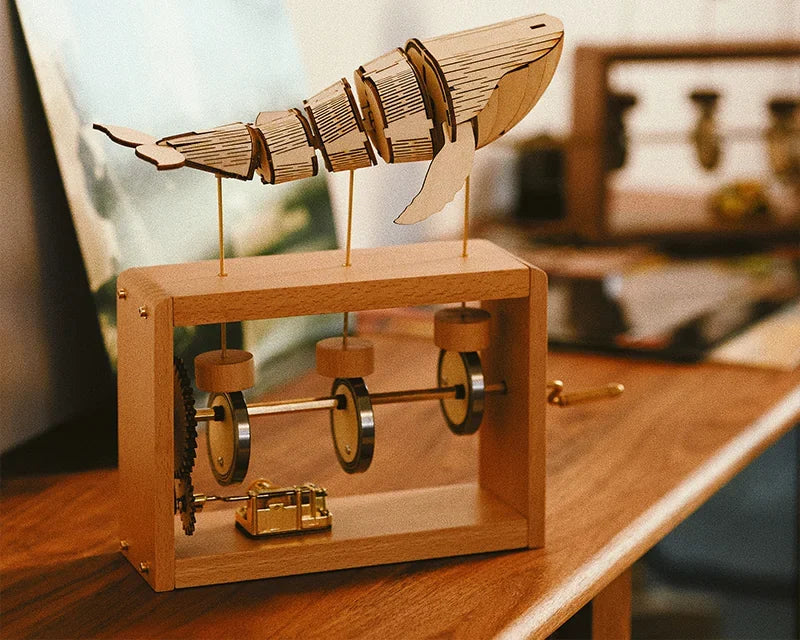 Light wood music box on a desk, a pale jointed whale on three rods above three round cams, a gold music box movement at the bottom of the frame.