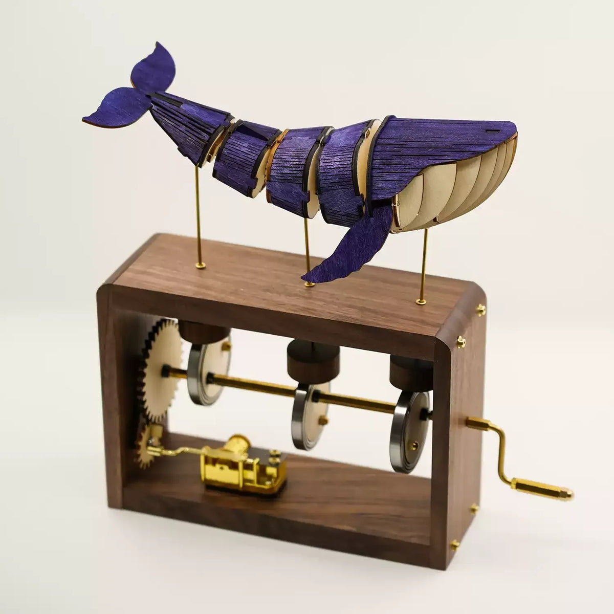 Walnut edition on white: a blue-violet jointed wooden whale on three brass rods above a dark walnut frame with gears, cams and a gold music box.