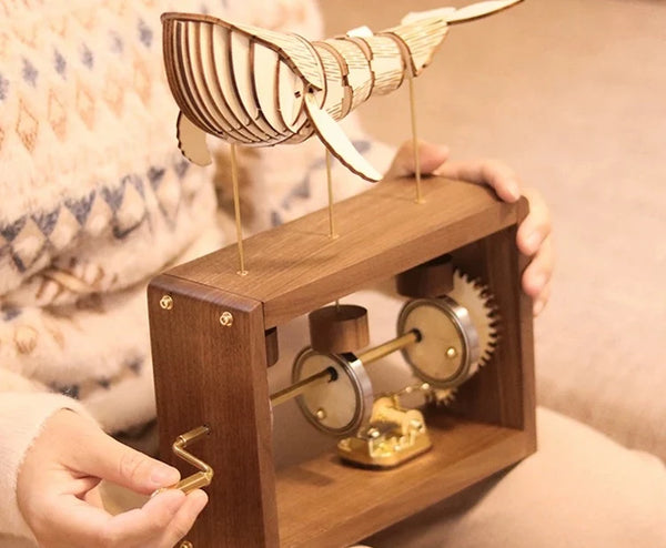 Two hands hold the walnut music box and turn its brass crank, a pale jointed wooden whale on brass rods above, a knit sweater behind.