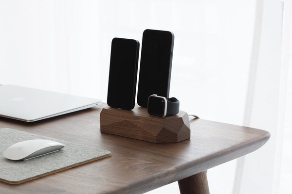 Walnut dock with two phones and a watch on a wooden desk beside a mouse and a felt mat