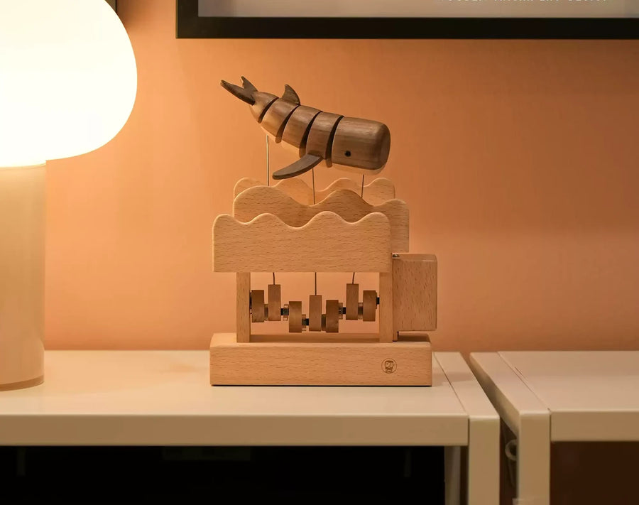 The whale automaton on a white shelf under a round lamp, its wave-topped box and cams in view.