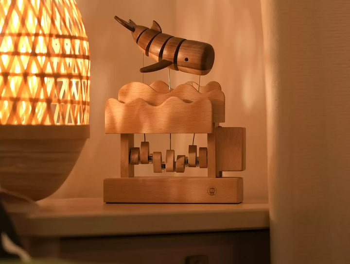 Striped wooden whale on fine wires above a wave-topped pale wood box with small cams inside, a woven lamp glowing beside it.